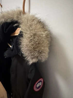 Svart dunjacka från Canada Goose XS - Säljer en svart dunjacka från Canada Goose i storlek XS. Jackan har en stor huva med beige pälsdetalj, dragkedja och knappar framtill samt två fickor. Perfekt för kalla vinterdagar och riktigt snygg streetstil.