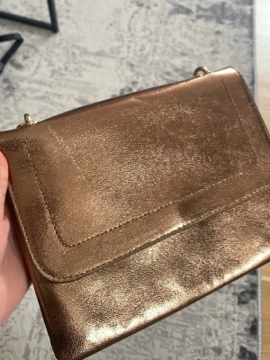 Guldig axelväska med kedjerem - Säljer en snygg axelväska i metallic guld med stilren design och kedjerem i matchande färg. Väskan har ett slätt yttre och praktiskt lock. Perfekt för dig som vill addera lite extra bling till din outfit.