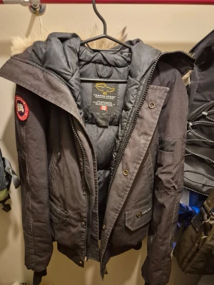Svart Canada Goose dunjacka med päls - Säljer en svart dunjacka från Canada Goose med ikonisk patch på ärmen och stor huva med beige päls. Jackan har dragkedja, knappar och flera fickor. Perfekt för kalla vinterdagar och riktigt varm. Klassisk och tidlös modell.