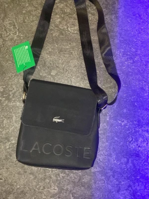Svart axelväska från Lacoste - Snygg svart axelväska från Lacoste med justerbar rem och präglad logga på framsidan. Väskan har magnetlås, ett huvudfack och en dragkedjeficka på baksidan. Tillverkad i slitstark syntet med diskreta detaljer och stilren design.