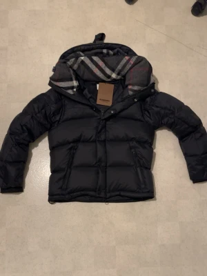 Svart Burberry dunjacka med avtagbara ärmar - Svart dunjacka från Burberry med avtagbara ärmar, så du kan bära den som väst. Jackan har klassiskt rutigt foder i huvan, dragkedja och knappar framtill samt två sidofickor med dragkedja. Perfekt för kalla dagar och riktigt snygg stil.