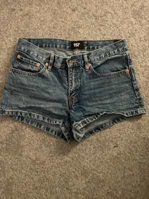 Blå jeansshorts från 157 - Säljer ett par klassiska blå jeansshorts från 157 med fem fickor, bälteshällor och knappgylf. Shortsen har en rak passform och är tillverkade i slitstark bomull. Perfekta för varma dagar och enkla att matcha med allt.