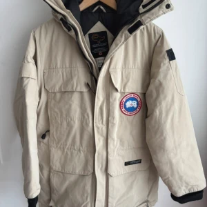 Beige Canada Goose dunjacka XXS - Säljer en beige äkta Canada Goose dunjacka med huva och stora fickor framtill. Jackan har klassisk patch på bröstet och dragkedja. Perfekt för kalla vinterdagar och riktigt snygg till streetwear-looken. Storlek är xxs men motsvarar s/m 