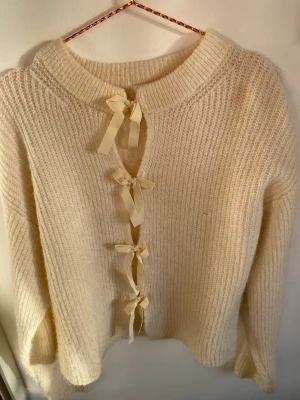 Beige stickad kofta med rosetter Gina Tricot - Supersöt beige stickad kofta från Gina Tricot med fyra dekorativa rosetter framtill istället för knappar. Koftan har långärmad passform och är gjord i ett mjukt, fluffigt material som känns riktigt mysigt. 