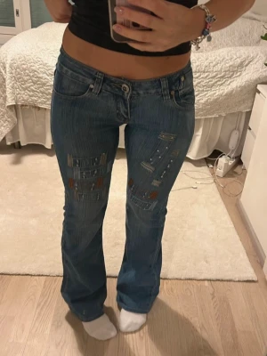 Low waist bootcut jeans med coola detaljer - Säljer ett par blå jeans med låg midja och bootcut💕Jeansen har unika paljettdetaljer på bakfickorna och coola slitningar med broderade inslag framtill. Köpte jeansen från Vinted men de var försmå för mig. Passar på 160-164 cm lång och midjemått är 35cm. Har inga tecken. Obs! Inte mina bilder. 