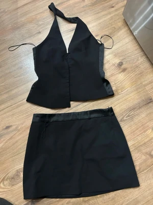 Svart set med satin-detaljer - Säljer en svart kortkjol med stilrena satin-detaljer i midjan. Kjolen har en enkel och clean look, perfekt för dig som gillar minimalistisk stil. Materialet känns mjukt och har en lätt glans. Kjolen har dragkedja i sidan för enkel påklädning.