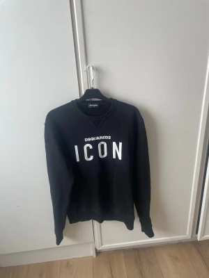 Svart sweatshirt Dsquared2 ICON - Svart sweatshirt från Dsquared2 med stor vit 'ICON'-text på bröstet och rund hals. Tröjan har långa ärmar och ribbade muddar vid ärmslut och nederkant. Tillverkad i mjuk bomull med lite stretch för extra komfort. Perfekt för dig som gillar streetwear och snygga loggor.