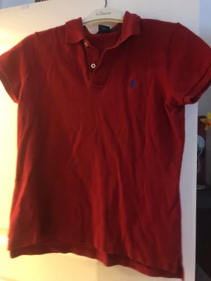 Röd pikétröja från Ralph Lauren - Säljer en snygg röd pikétröja från Ralph Lauren, modell The Skinny Polo. Tröjan har klassisk krage, korta ärmar och två knappar framtill. Materialet är 100% bomull och det finns en liten blå broderad logga på bröstet. Perfekt för en chill och stilren look.