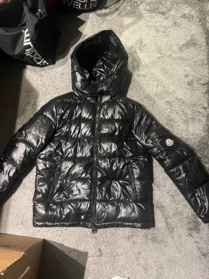 Svart Moncler pufferjacka med huva - Säljer en svart pufferjacka från Moncler med glansig finish och huva. Jackan har dragkedja framtill, två sidofickor med dragkedja och Moncler-logga på ärmen. Perfekt för kalla dagar och riktigt snygg till streetwear. Använd en vinter, köpte den ny. Storlek 1, kom privat för flera bilder