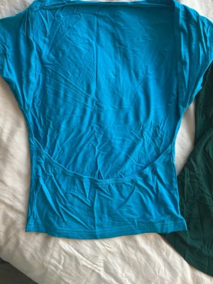 toppar med öppen rygg - Säljer två enkla och mjuka toppar i stretchigt material, en i klarblå och en i mörkgrön färg. Båda har rund halsringning och är kortärmade med en figurnära passform. Perfekta att matcha till jeans eller kjol. Öppna i ryggen