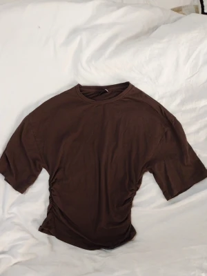 Brun topp med rynkade sidor Gina Tricot - Säljer en brun topp  från Gina Tricot i storlek XS. T-shirten har korta ärmar och rynkade sidor. Sparsamt använd och i bra skick! Katt finns I hemmet.