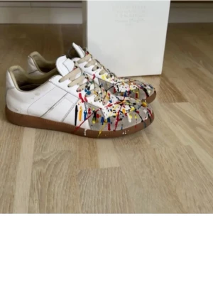 Maison Margiela sneakers med färgstänk - Maison Margiela sneakers i vitt skinn med beige detaljer och coola färgstänk i rött, blått, gult och svart. Klassisk låg modell med snörning och brun sula. Unik design som sticker ut och passar perfekt till streetwear eller casual outfits.
