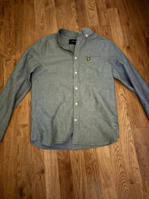 Grå blå skjorta från Lyle & Scott - Säljer en stilren grå skjorta från Lyle & Scott med klassisk krage och knappstängning. Skjortan har en bröstficka med den ikoniska gula loggan broderad. Tillverkad i mjuk bomull och har långa ärmar. Perfekt till jeans eller chinos.