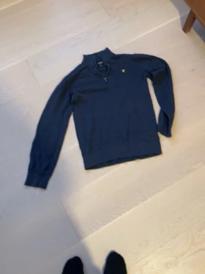 Marinblå half zip tröja Lyle & Scott - Snygg marinblå långärmad tröja från Lyle & Scott Junior med half zip och gul logga på bröstet. Tröjan har hög krage och är tillverkad i mjukt material, perfekt för kyligare dagar.