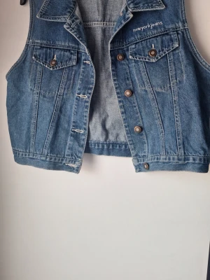 Blå jeansväst från NY Jeans - Säljer en klassisk blå jeansväst från NY Jeans i 100% bomull. Västen har två bröstfickor med knappar, snygga sömdetaljer och metallknappar framtill. Perfekt att slänga över en t-shirt för en cool och avslappnad look.