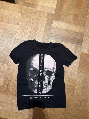 Svart Philipp Plein t-shirt med döskalle - Snygg svart t-shirt från Philipp Plein med stor vit döskallegrafik på framsidan och texten 'ORIGINALS' nedtill. T-shirten har rund hals och korta ärmar, perfekt för dig som gillar edgy streetwear.