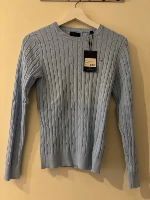 Ljusblå Gant tröja  - Snygg ljusblå kabelstickad tröja från GANT. Tröjan har rund hals, ribbade muddar och lång ärm. Liten GANT-logga broderad på bröstet.