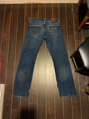 Levi's 506 Standard blå jeans - Klassiska Levi's 506 Standard jeans i mörkblå tvätt med snyggt slitna detaljer. Rak passform, fem fickor och ikoniska röda Levi's-lappen bak. Tillverkade i slitstarkt denim med kontrastsömmar och normal midja. Perfekta för en avslappnad och tidlös look.