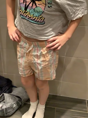 Randiga pastellshorts från Lindex och en mjukis tröja från HM - Säljer ett par mjuka och lätta shorts  och tröja från Lindex och hm  i storlek 134/140.  Shortsen har ett pastellfärgat rutmönster i persika, ljusblått och rosa, samt elastisk midja med vit snörning. Perfekta för varma dagar och chill häng.