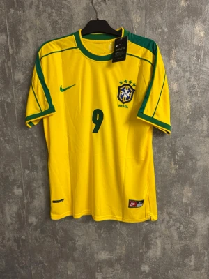 Nike Brasil Ronaldo 9 fotbollströja - XL - Gul Brasilien fotbollströja från Nike med gröna detaljer, nummer 9 och Ronaldo på ryggen. Broderat CBF-emblem på bröstet och Nike-logga. Tillverkad i polyester med Dri-FIT och Breathe-teknologi för extra komfort. Perfekt för dig som älskar fotboll och Brasilien.