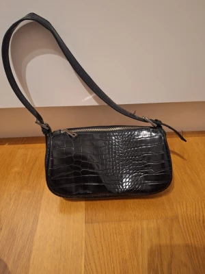 Svart vintage gandväska från Gina Tricot - Snygg svart axelväska från Gina Tricot med croco-mönstrad yta och silverfärgad dragkedja. Väskan har en stilren, avlång form och ett justerbart axelband.  Väldigt rymlig. Cool vintage väska. 