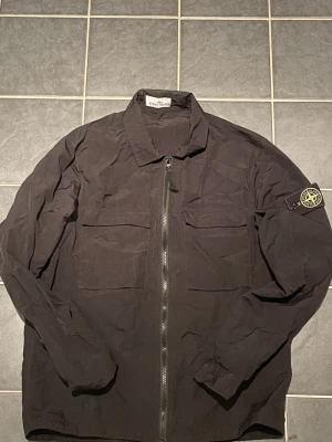 Svart overshirt från Stone Island - Svart overshirt från Stone Island med klassisk krage, två stora bröstfickor och dragkedja framtill. Ikonisk patch på vänster ärm. Perfekt för lager-på-lager och har en clean, modern look som funkar till många stilar.