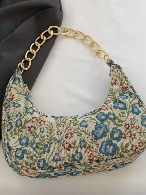 Blommig axelväska med guldkedja - Säljer en snygg axelväska med blommigt mönster i blått, grönt och rött på beige bakgrund. Väskan har en chunky guldfärgad kedja som axelrem och dragkedja upptill. Helt ny, hör av dig vid frågor 🥰