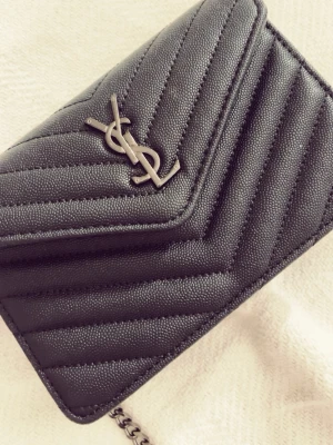YSL väska - Säljer en svart quiltad axelväska från YSL med klassisk silverfärgad logga framtill. Väskan har kedjerem och ett snyggt mönster med diagonala sömmar. Insidan är vinröd med dragkedjefack för småsaker. Perfekt storlek för det viktigaste.