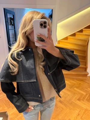 Mörkblå jeansjacka med cape-detalj - Säljer en riktigt snygg mörkblå jeansjacka med cool cape-liknande detalj över axlarna. Jackan har markerade sömmar, tryckknappar framtill och stora fickor. Perfekt för dig som vill sticka ut lite extra i vår eller höst.