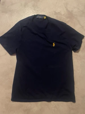 Mörkblå t-shirt från Polo Ralph Lauren - Snygg mörkblå t-shirt från Polo Ralph Lauren med klassisk gul broderad logga på bröstet. T-shirten har rund hals och korta ärmar, perfekt för en clean och stilren look. Tillverkad i mjuk bomull för skön känsla hela dagen.