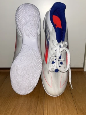 Adidas F50 vita och blå fotbollsskor - Säljer ett par Adidas F50 fotbollsskor i vitt med blå och röda detaljer. Skorna har snörning, låg profil och platt sula för inomhusspel. Ovandelen är i syntetmaterial med klassiska Adidas-ränder på sidan och F50-logga på plösen och innersulan. Perfekta för dig som vill sticka ut på planen. Dom är också oanvända. Tyvärr har dock ingen kartong, kostar 700 säljer för 499,skriv om du har frågor 