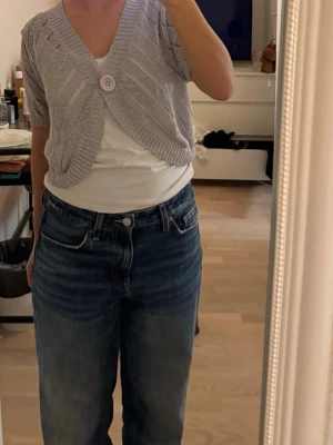 Ljusgrå kort kofta med knapp - Säljer en ljusgrå, kort kofta med v-ringning och en stor dekorativ knapp framtill. Koftan har ett stickat mönster med diskreta hål och ribbade kanter. 