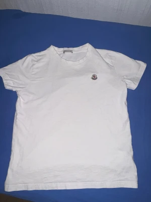 Vit Moncler t-shirt  - Säljer en clean vit t-shirt från Moncler med klassisk rund hals och diskret logga på bröstet. Tillverkad i mjuk bomull som känns skön mot huden. Perfekt till jeans eller shorts för en enkel men snygg look.