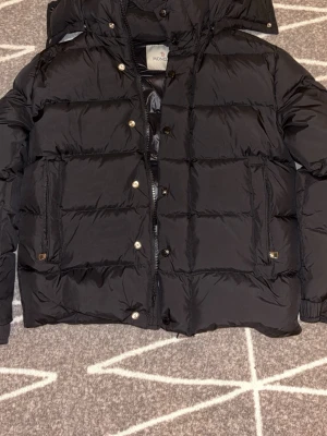 Svart Moncler pufferjacka med huva - Säljer en svart pufferjacka från Moncler med huva och klassisk logga på ärmen. Jackan har dragkedja och knappar framtill, två stora fickor och quiltad design. Materialet är glansig polyester och dunfyllning som håller dig varm under kalla dagar. PRIS KAN DISKUTERAS 