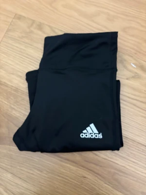 Svarta Adidas tights med vita ränder - Säljer ett par svarta Adidas tights i storlek S. De har klassiska vita ränder längs ena benet och Adidas-logga på låret. Tillverkade i stretchigt syntetmaterial som passar perfekt till träning eller chill. Supersnygga och bekväma tights med hög midja.