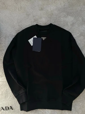 Svart sweatshirt från Prada - Säljer en svart sweatshirt från Prada med klassisk rund hals och diskret metallogo framtill. Tröjan har långa ärmar och ribbade muddar vid ärmslut och nederkant. Tillverkad i mjuk bomull för en clean och stilren look.