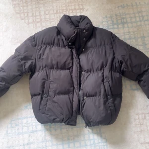 Svart pufferjacka med dragkedja - Säljer en svart pufferjacka med hög krage och dragkedja framtill. Jackan är quiltad och har två fickor med knappstängning. Perfekt för kalla dagar och har en oversized vibe. Passar dig som gillar en enkel men trendig look.