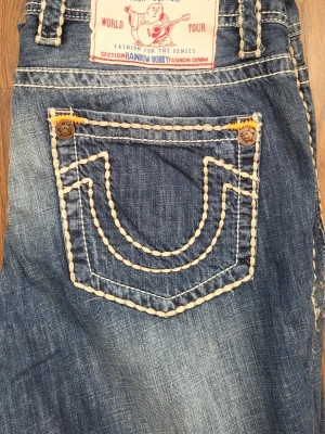 True Religion jeans med kontrastsömmar - Säljer ett par klassiska blå True Religion jeans med tydliga vita kontrastsömmar och ikoniska bakfickor. Jeansen har guldiga knappar, låg midja och slitna detaljer för en cool vintagekänsla. Tillverkade i USA i robust denim.