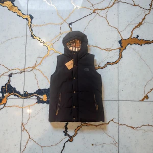 Svart dunväst med huva från Burberry - Svart dunväst från Burberry med huva och klassiskt rutigt foder. Västen har dragkedjor på fickorna, tryckknappar framtill och en quiltad look. Perfekt för lager-på-lager och snygg till streetwear.