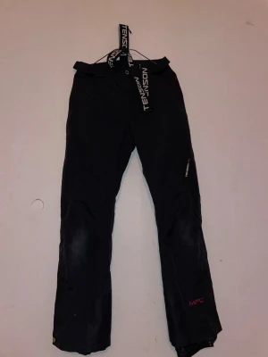 Svarta täckbyxor från Tenson - Svarta täckbyxor från Tenson med justerbara hängslen och knappstängning i midjan. Byxorna är tillverkade i polyester och har broderad logga på benet. Perfekta för vintern och outdoor-aktiviteter. Snygg och funktionell design med MPC-tryck på benet.