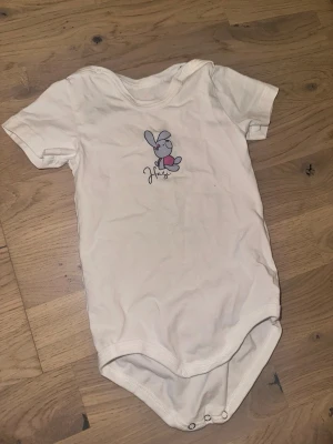 Vit body med kanintryck från Kanz Kanz vit body, storlek 74 / 9M - Söt vit body från Kanz i storlek 74/9M med korta ärmar och tryckknappar i grenen. Framtill finns ett gulligt tryck med en tecknad kanin och texten 'Hasi'. Perfekt för små barn som gillar mjuka och bekväma kläder.Vit body med korta ärmar och tryck av en kanin med ett hjärta. 