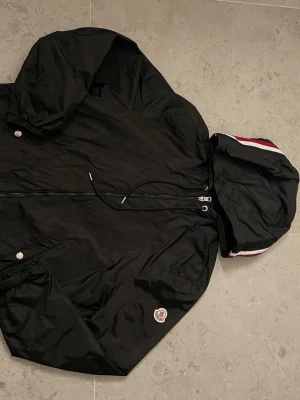 Svart Moncler vindjacka med huva - Svart vindjacka från Moncler med huva och snygga rödvita detaljer på kanten. Jackan har dragkedja framtill, två fickor med tryckknappar och Moncler-logga på ärmen. Materialet är lätt och känns sportigt, perfekt för dig som gillar stilren design.