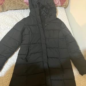 Svart lång pufferjacka från 157 - Säljer en svart pufferjacka från 157 i storlek M. Jackan är lång och har huva. Perfekt för kalla dagar och har dragkedja framtill. I mycket bra skick, som ny.