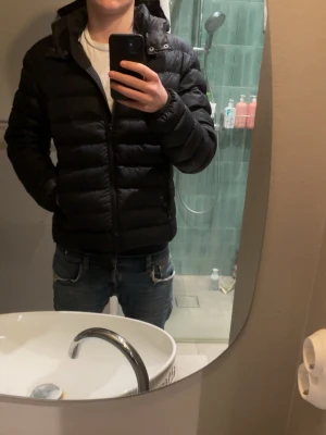 Svart Colmar jacka herr - Svart pufferjacka från Colmar Originals med huva och dragkedja framtill. Jackan har ett stilrent, quiltat mönster och logotyp på ärmen. Perfekt för kalla dagar och har en snygg, normal passform. Materialet är polyester och dunfyllning. Köpt i butik och använd fåtal gånger. STORLEK S (50)