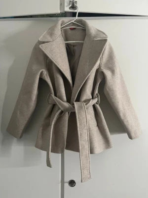 Beige kappa med bälte och krage - Snygg beige kappa med bred krage och bälte i midjan. Jackan har en klassisk omlottstil och är tillverkad i ett mjukt ull-liknande material. Perfekt för dig som vill ha en stilren och trendig look under kyliga dagar.