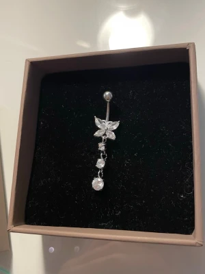 Silver navelpiercing med fjäril Montini - Säljer en snygg, ny navelpiercing från Ur&penn med hängande fjäril och glittrande stenar. Piercingen är i kirurgiskt stål och har en barbell-design med en kula upptill. Perfekt för dig som vill ha något extra till din look.