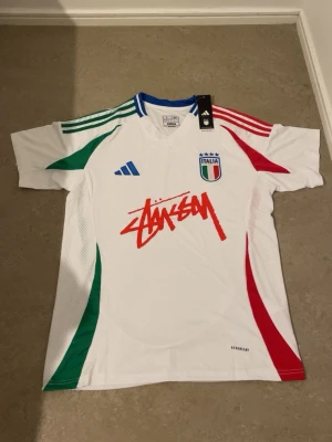 Vit Adidas fotbollströja Italien  - Säljer en vit fotbollströja från Adidas i storlek L, slim fit. Tröjan har gröna och röda detaljer, blå krage och Italiens landslagsmärke på bröstet. Stüssy-logga i rött framtill och Aeroready-material som andas. Perfekt för dig som gillar streetwear och fotboll.
