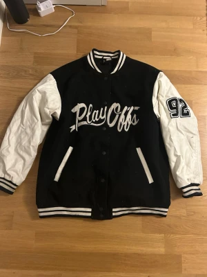 Svartvit bomberjacka  - Säljer en svartvit bomberjacka från Divided i storlek S. Jackan har vita ärmar, svart kropp och broderad text 'Play Offs' samt siffran 92 på ena ärmen. Klassiska ränder på muddar och krage, stängs med knappar framtill. Perfekt för en sportig och trendig look.
