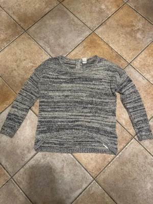 Gråmelerad stickad tröja Vero Moda - Mysig gråmelerad stickad tröja från Vero Moda i storlek S. Tröjan har ett randigt mönster i olika grå och mörkblå toner, rund halsringning och lång ärm. Dragkedja bak i nacken ger en snygg detalj. Perfekt för kyliga dagar!