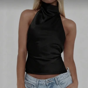 Svart omlott halterneck topp - Säljer en svart halterneck topp med omlott-design och öppen rygg. Toppen har en snygg rosett i nacken och smala band som knyts runt ryggen. Materialet är glansigt och känns lyxigt, perfekt för att lyfta din outfit.